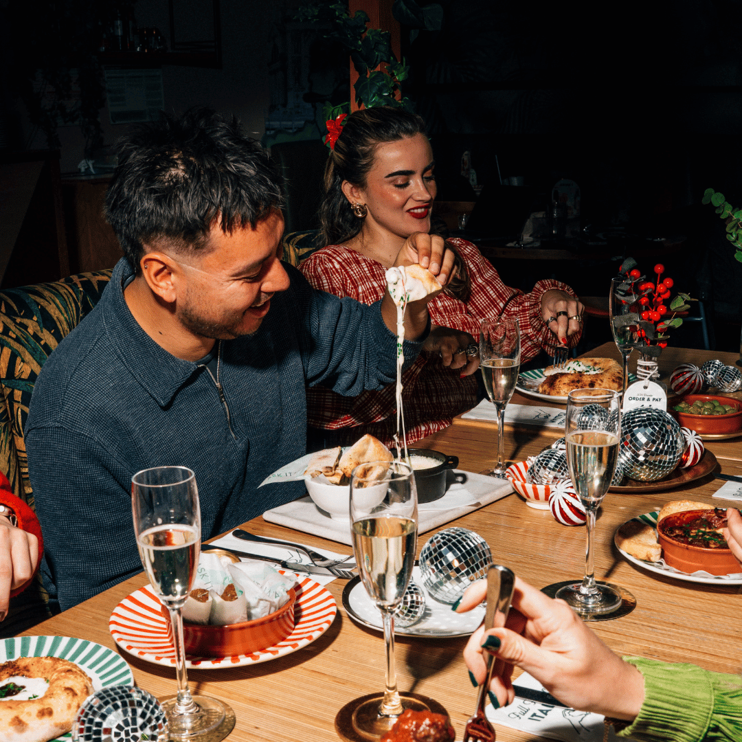 Photo of somebody sat round a festive table at a restaurant, laughing at the cheese pull from a cheese fondue. 